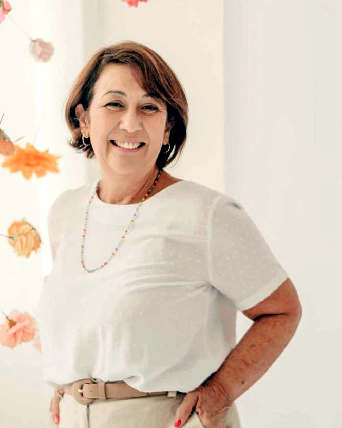 Vera Lima Portes, sorrindo, de blusa branca, diante de uma parede com flores
